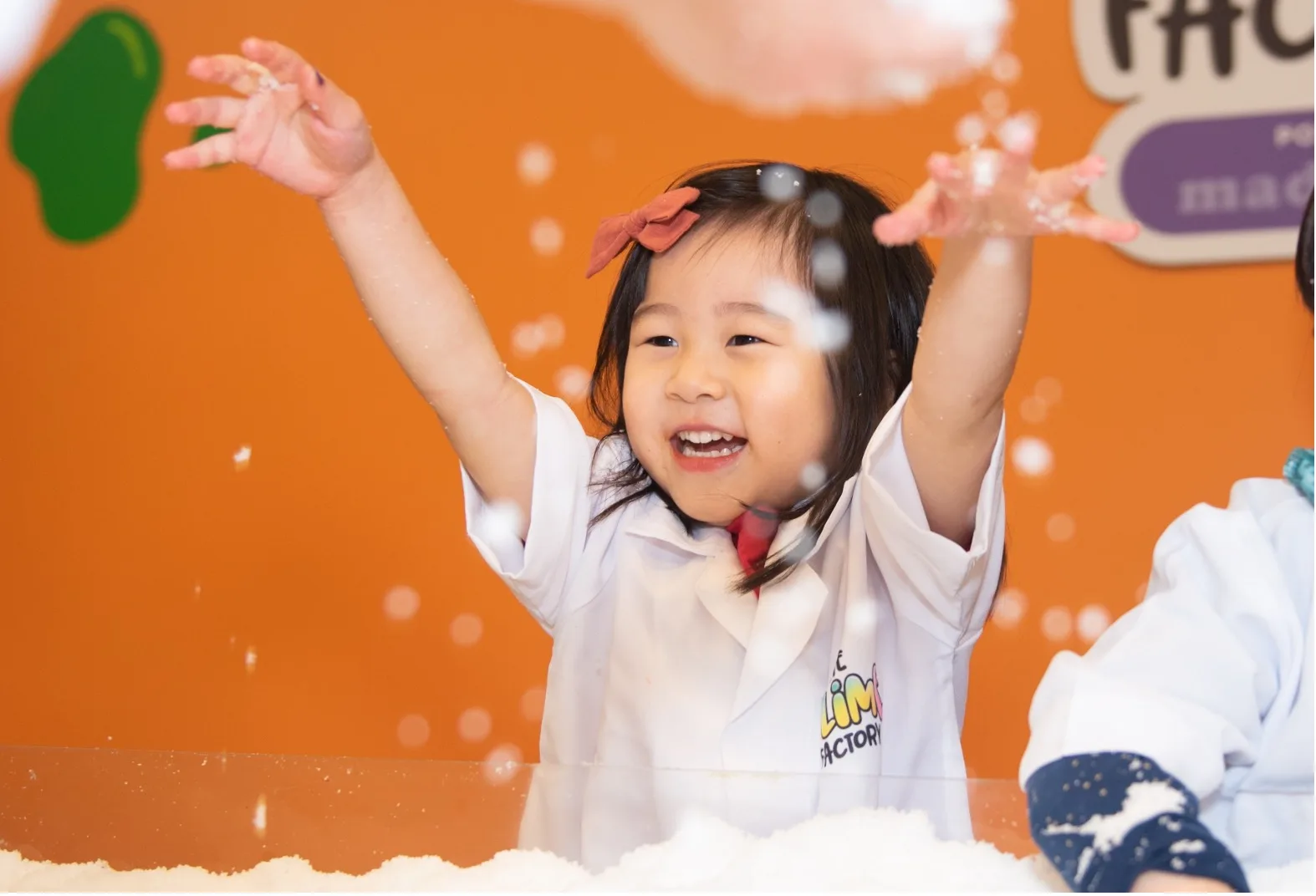 A young guest raises her hands while making slime beside the orange Slime Factory wall in Bellevue.