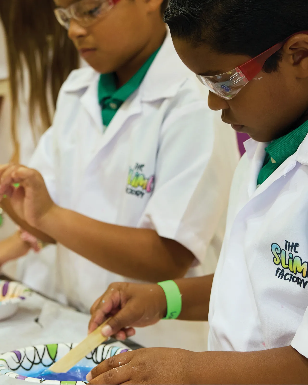 Young scientists wearing lab coats and goggles mix colorful slime at the lab bench.