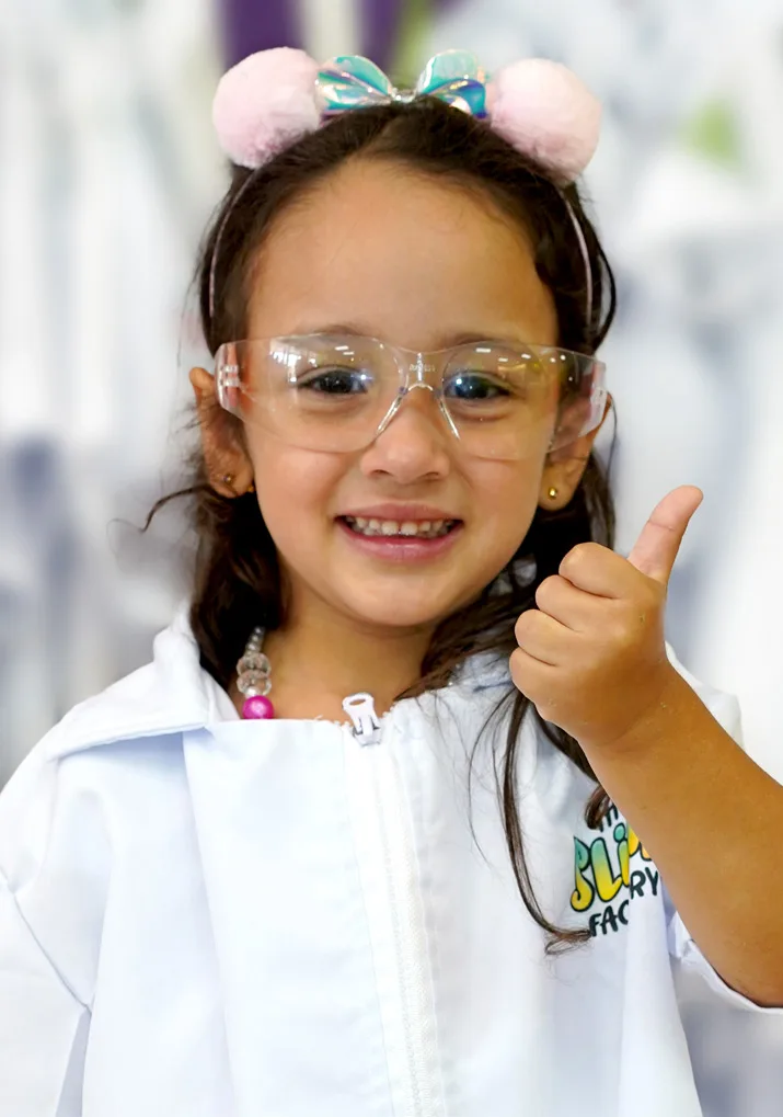 A smiling young scientist wearing safety goggles and a white lab coat gives a thumbs-up.