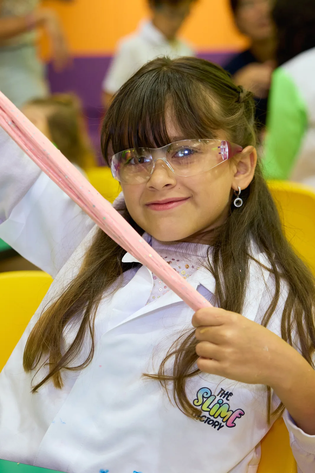 A smiling young scientist wearing goggles and a lab coat stretches a ribbon of pink slime.