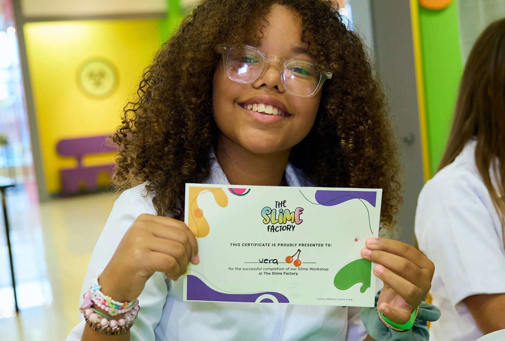 A smiling guest wearing safety goggles and a lab coat holds a Slime Factory workshop certificate.