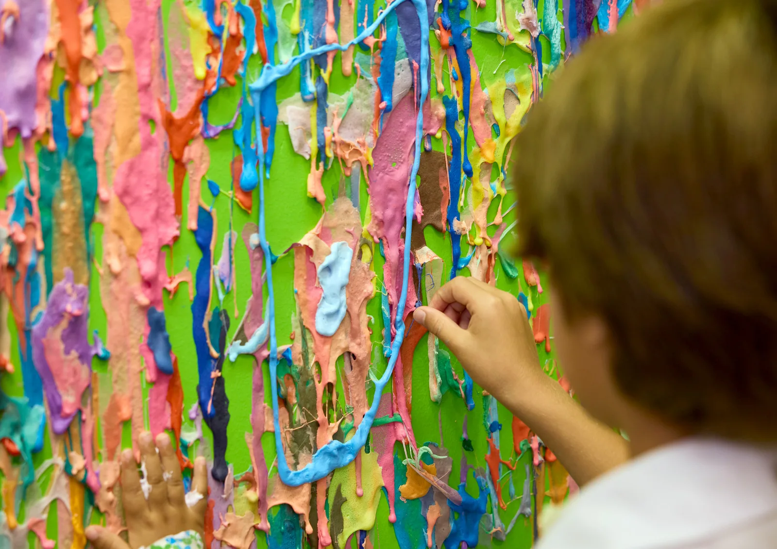 A young scientist adds colorful slime to a wall covered in bright slime creations.